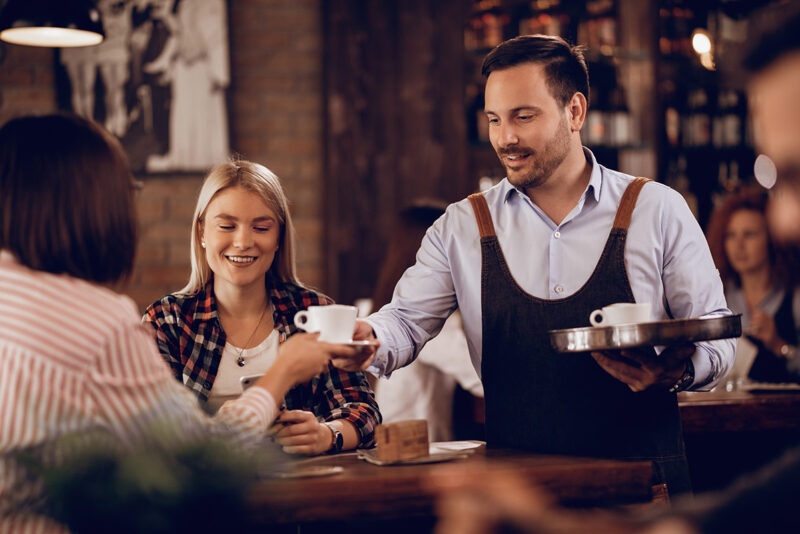 smiling-waiter-serving-coffee-female-guests-cafe